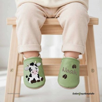 Lederpuschen in pistazie-creme mit Namen (cappuccino) und Hufabdrücken (dunkelbraun) links sowie rechts Kuh (weiß-schwarz-rosa-dunkelbraun. Designbeispiel Ki-generiert Baby auf Hocker