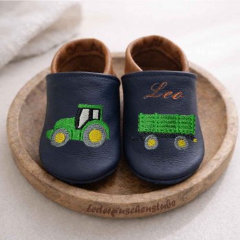 Handgefertigte Krabbelschuhe in dunkelblau-braun, Traktor und Anhänger mittelgrün-gelb links Namen braun Spann Designbeispiel auf einem Holzteller gestellt