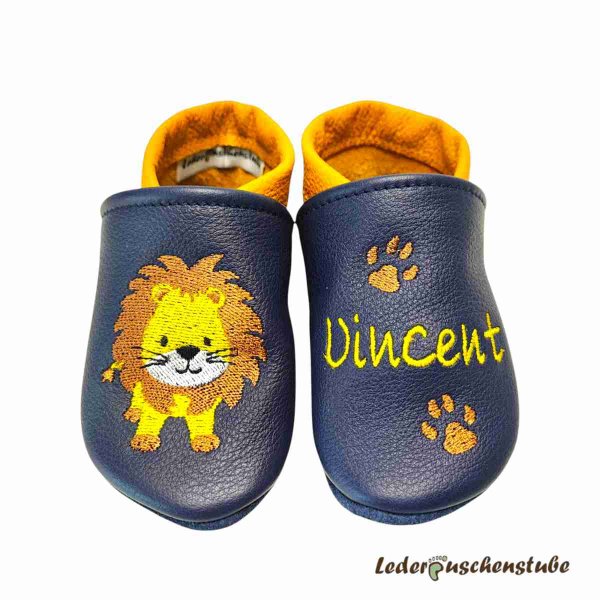 Krabbelschuhe in dunkelblau-maisgelb, rechts Löwe steht (gelb-braun-weiß-schwarz) und links Name (gelb) mit 2 Tatzen (braun)